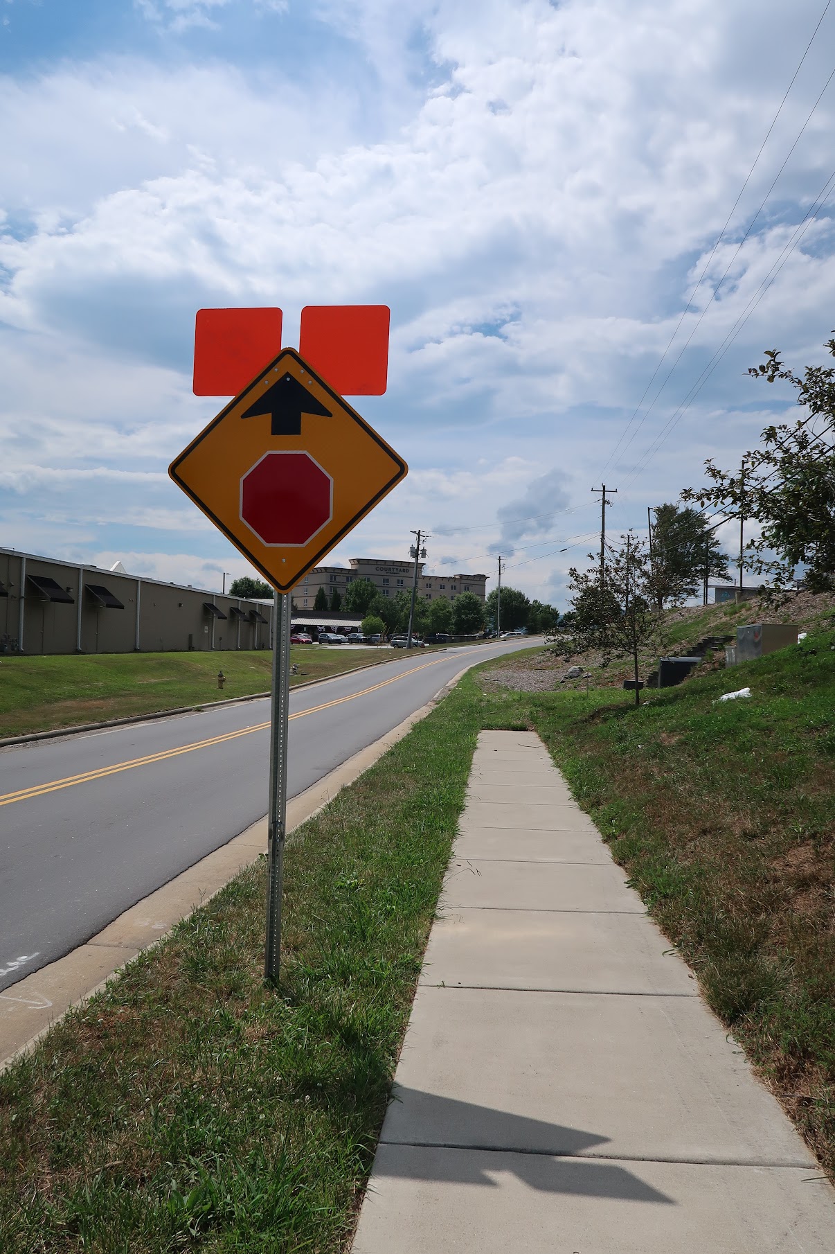 Traffic sign on a pole: yellow diamond with an up arrow and a red stop sign inset, two orange panels above near a sidewalk road.