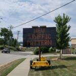 Mobile LED traffic sign on a trailer displaying a message about new traffic; utility poles and a sunny street in the background.