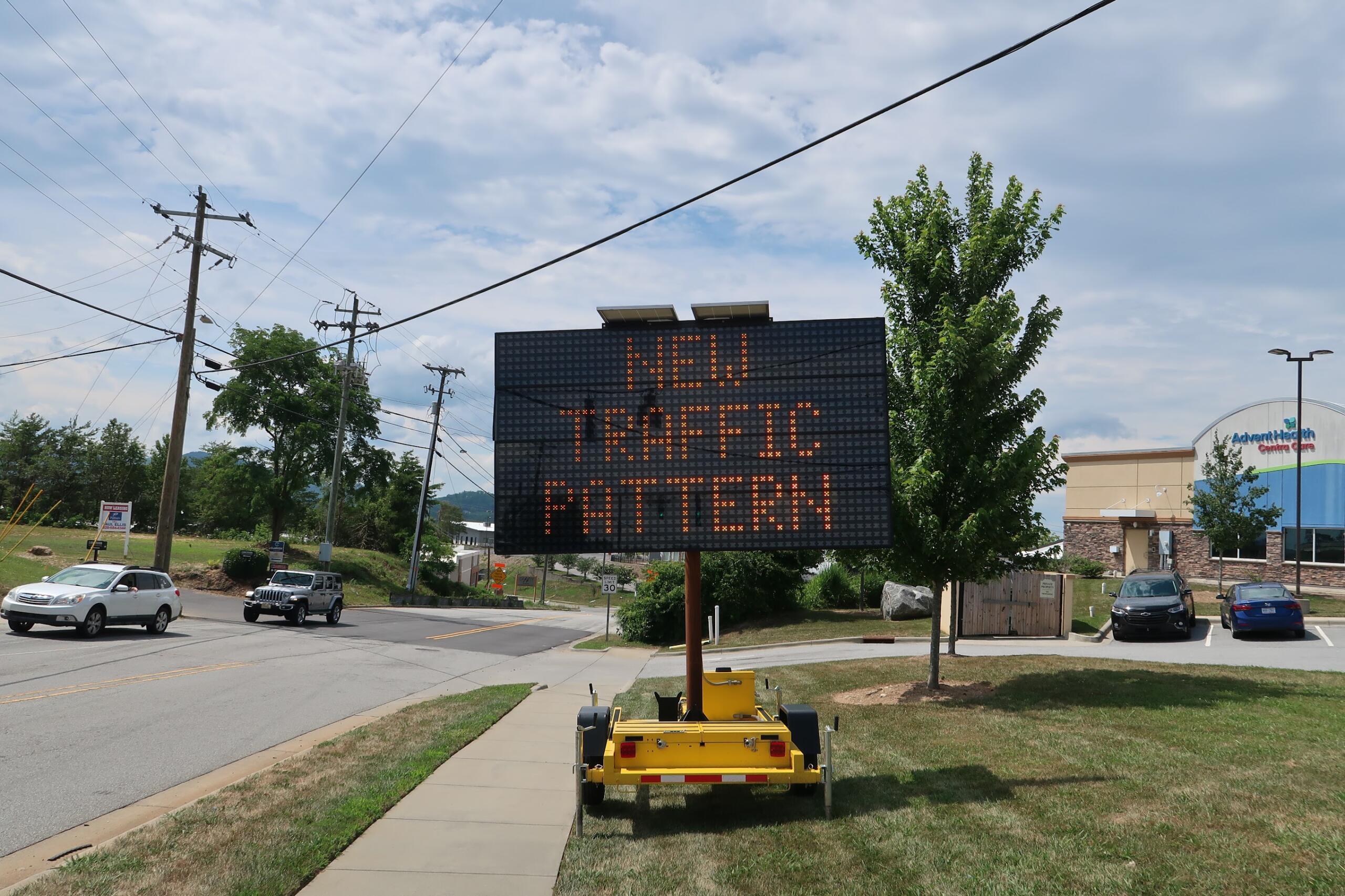 Mobile LED traffic sign on a trailer displaying a message about new traffic; utility poles and a sunny street in the background.