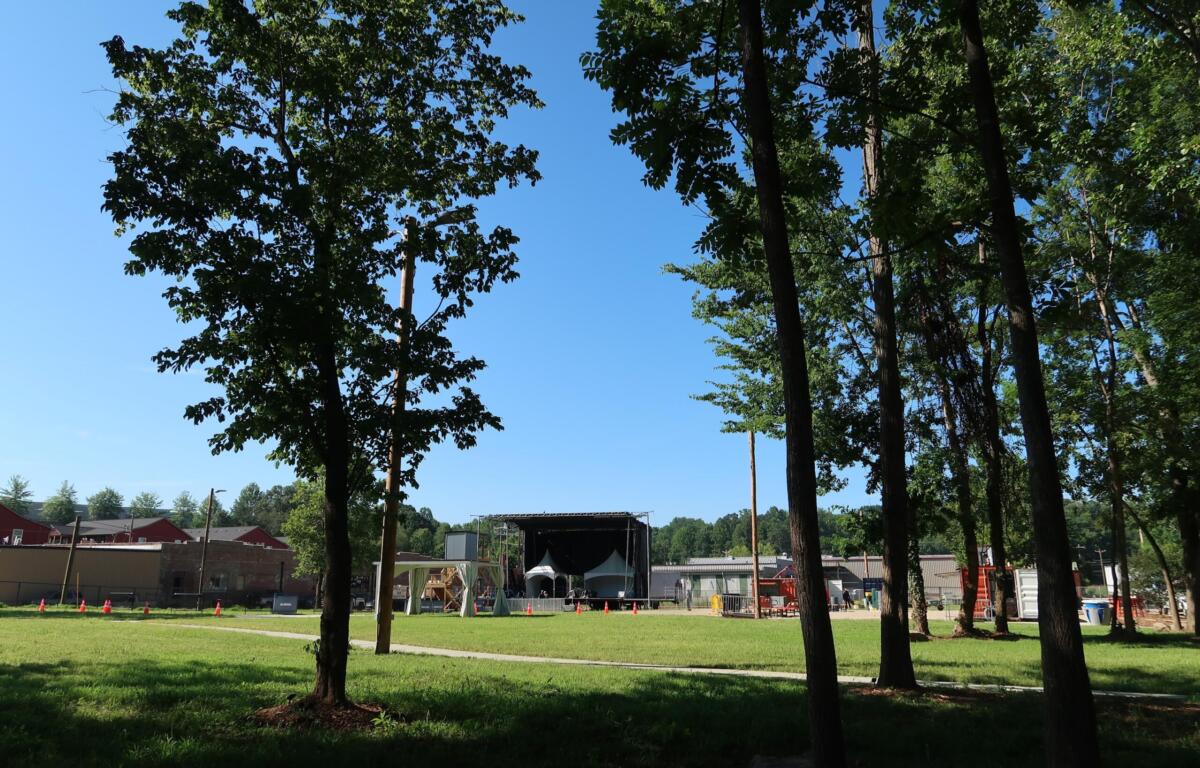 Open grassy park with tall leafy trees and a clear blue sky; a large stage and event tents are visible in the background.