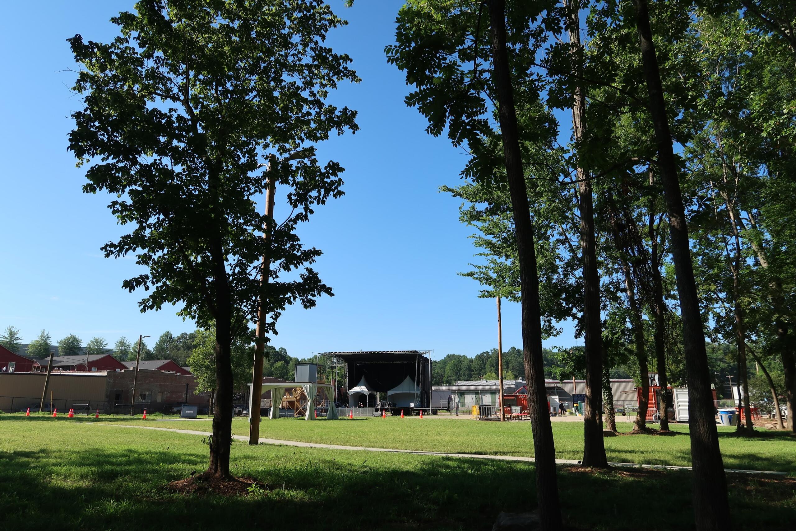 Open grassy park with tall leafy trees and a clear blue sky; a large stage and event tents are visible in the background.