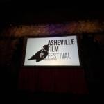 Sign at a film festival showing 'Asheville Film Festival' with a bear graphic on a rocky wall backdrop (dimly lit).