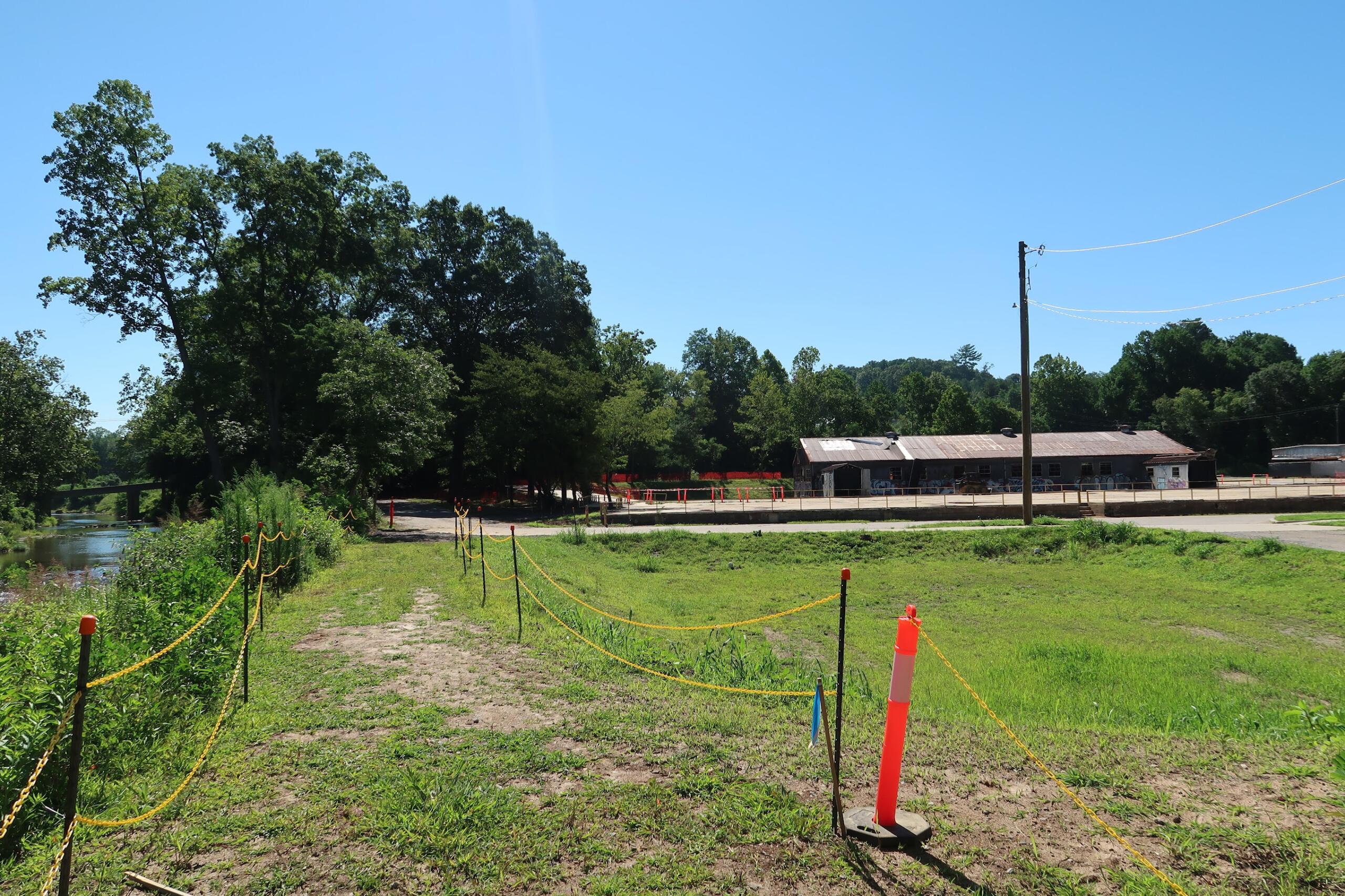 Sunny rural scene with a dirt path, yellow caution tape on posts, and orange cones leading toward a long, rusty-roofed building; trees and a river to the left under a clear blue sky.