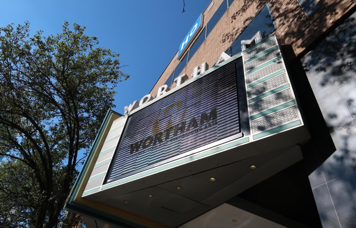 Low-angle view of a brick building with a large digital marquee showing the word 'WORLD' and glass panels against a blue sky with trees nearby.