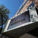 Low-angle view of a brick building with a large digital marquee showing the word 'WORLD' and glass panels against a blue sky with trees nearby.