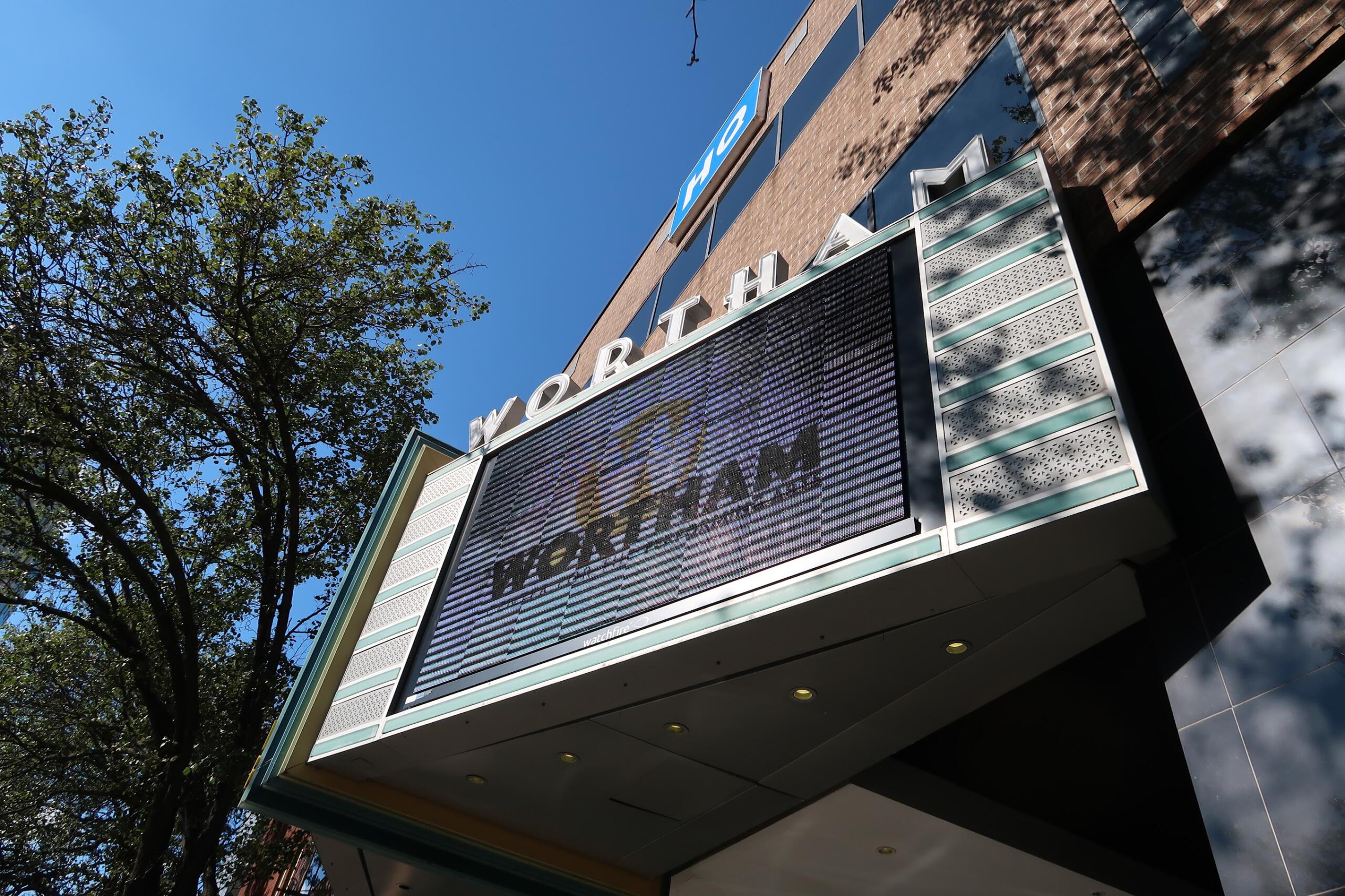 Low-angle view of a brick building with a large digital marquee showing the word 'WORLD' and glass panels against a blue sky with trees nearby.