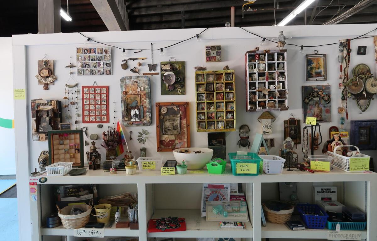 Shop display of handmade craft items: framed photos, jewelry, trinkets, and small boxes arranged on shelves with price tags and baskets beneath.