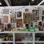 Shop display of handmade craft items: framed photos, jewelry, trinkets, and small boxes arranged on shelves with price tags and baskets beneath.