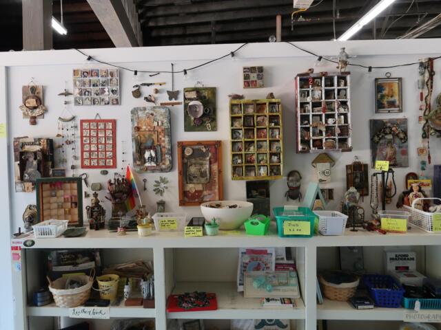 Shop display of handmade craft items: framed photos, jewelry, trinkets, and small boxes arranged on shelves with price tags and baskets beneath.
