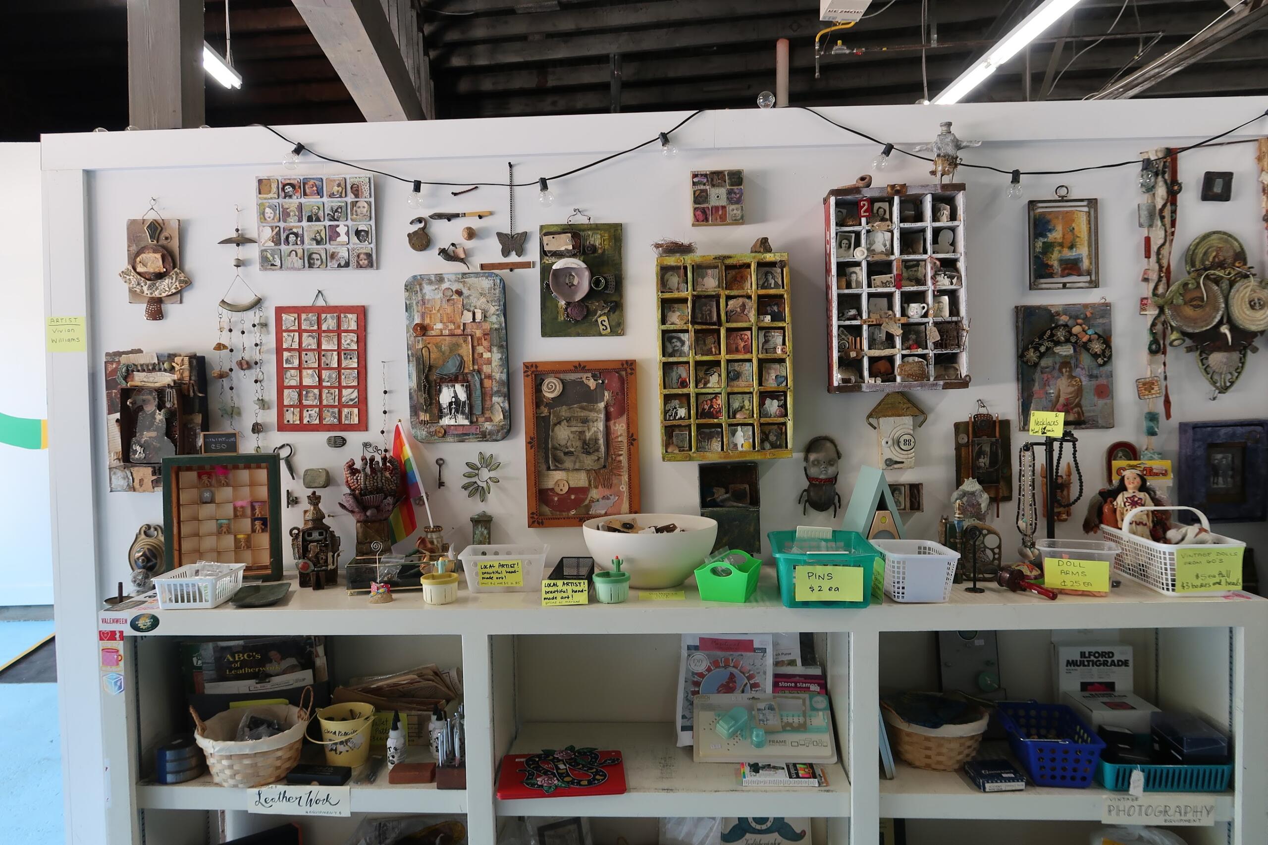 Shop display of handmade craft items: framed photos, jewelry, trinkets, and small boxes arranged on shelves with price tags and baskets beneath.