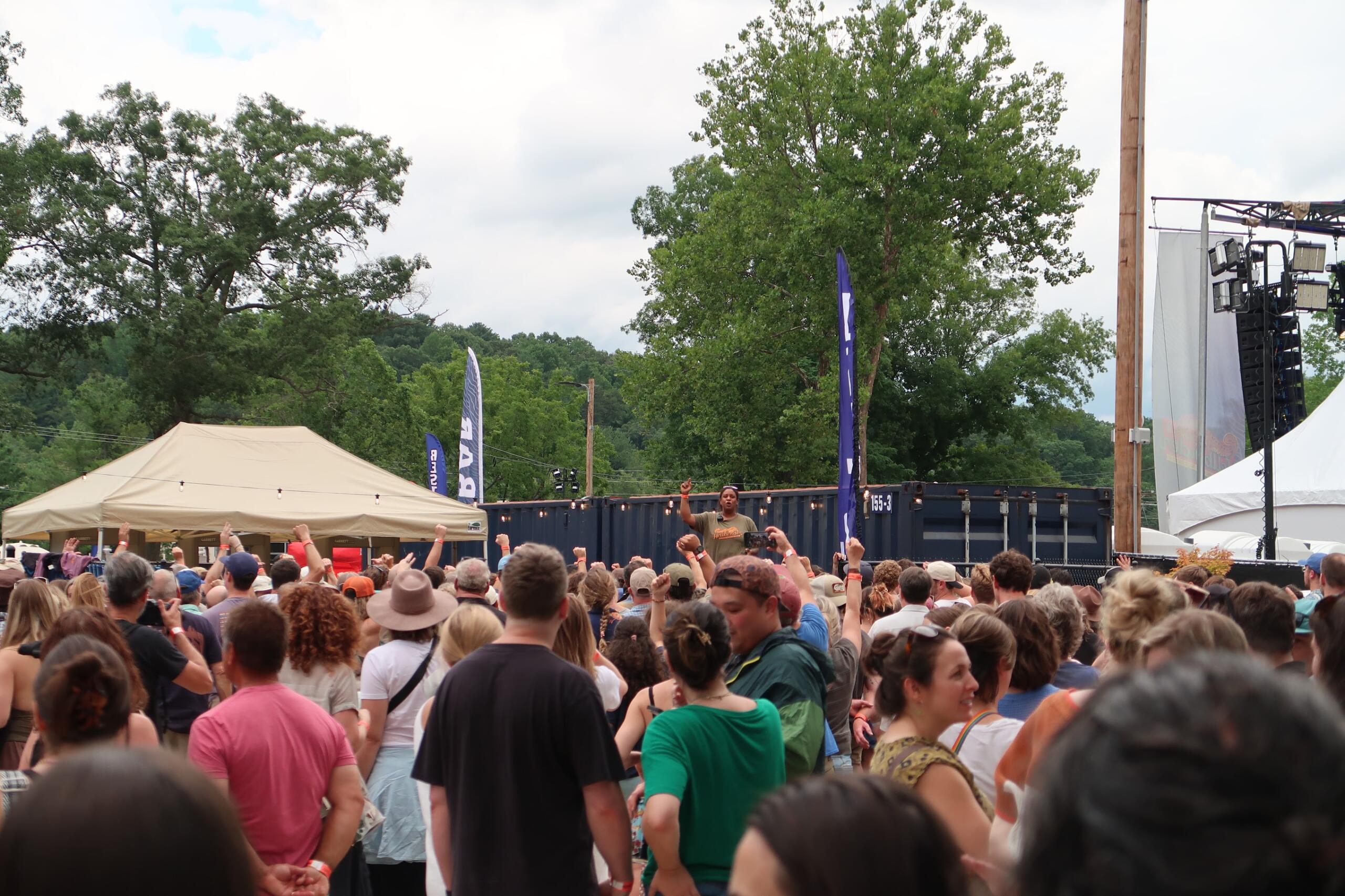 Large crowd at an outdoor concert, stage visible on the right with lighting rigs and a cloudy sky.