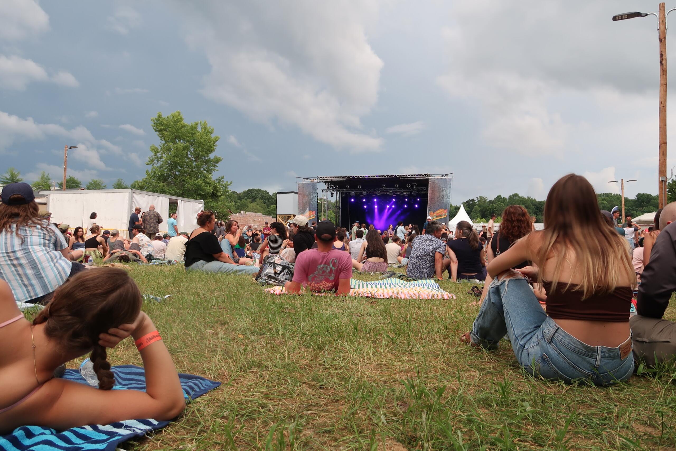 Crowd seated on grass at an outdoor concert, stage with purple lights in the distance.