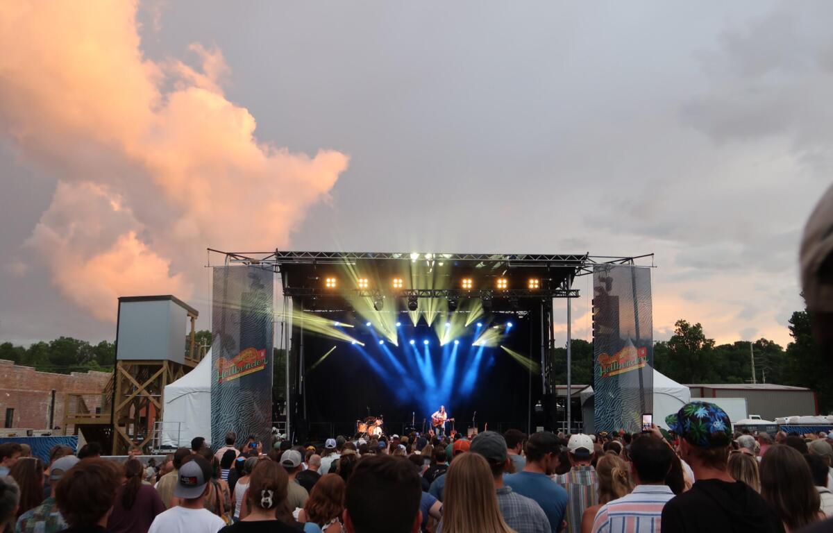 Outdoor music festival crowd facing a lit stage at dusk, with orange sunset clouds in the sky and bright stage lights.