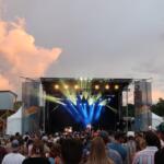 Outdoor music festival crowd facing a lit stage at dusk, with orange sunset clouds in the sky and bright stage lights.