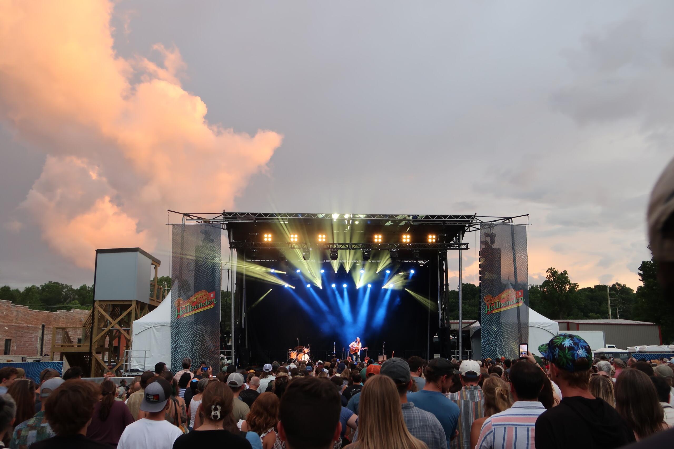 Outdoor music festival crowd facing a lit stage at dusk, with orange sunset clouds in the sky and bright stage lights.