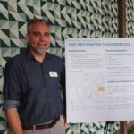 Man standing beside an informational poster on 'Tree Restoration Considerations,' indoors, patterned geometric wall in background.