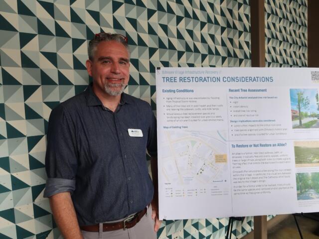 Man standing beside an informational poster on 'Tree Restoration Considerations,' indoors, patterned geometric wall in background.