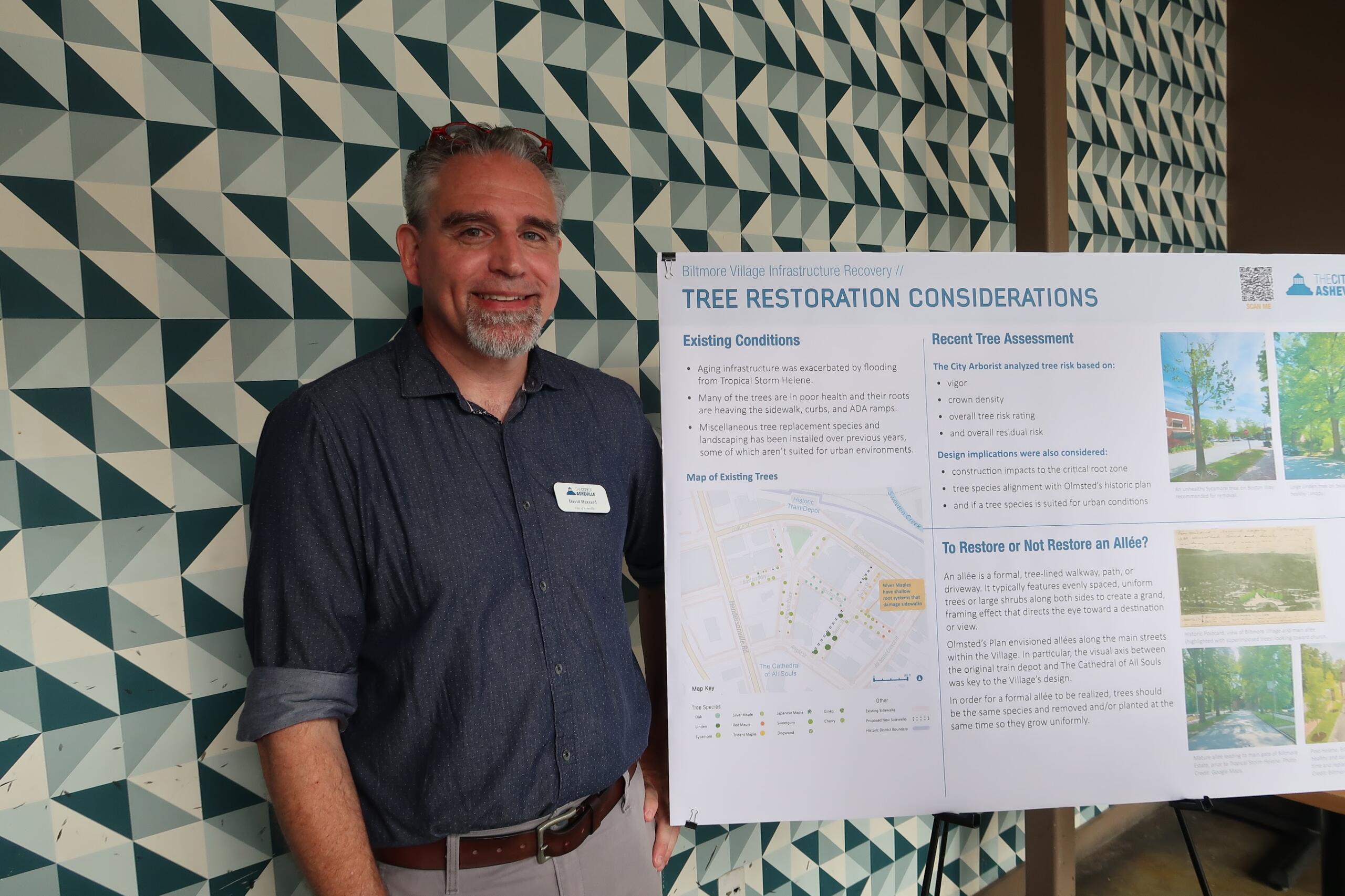 Man standing beside an informational poster on 'Tree Restoration Considerations,' indoors, patterned geometric wall in background.