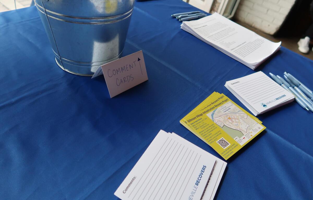 Table set up for feedback with a metal bucket, stacks of comment cards, pens, and printed sheets on a blue tablecloth.