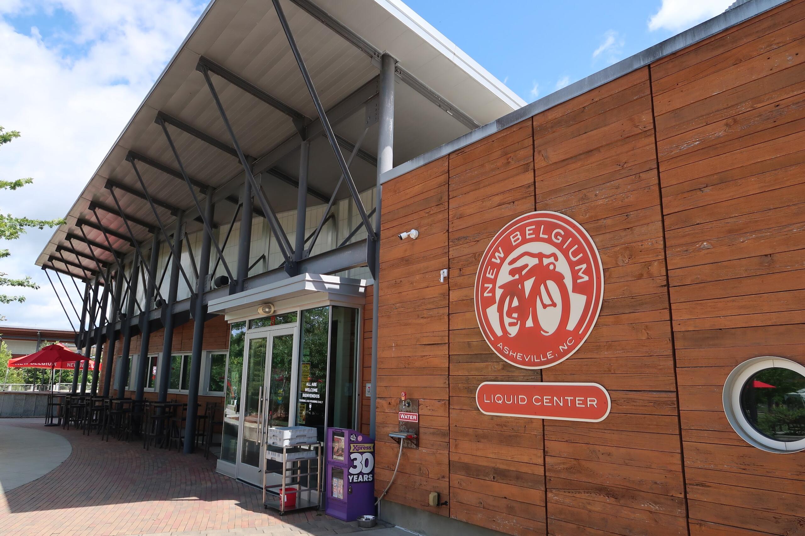 Exterior of a wood-clad building with a large circular New Belgium logo featuring a bicycle on the wall.
