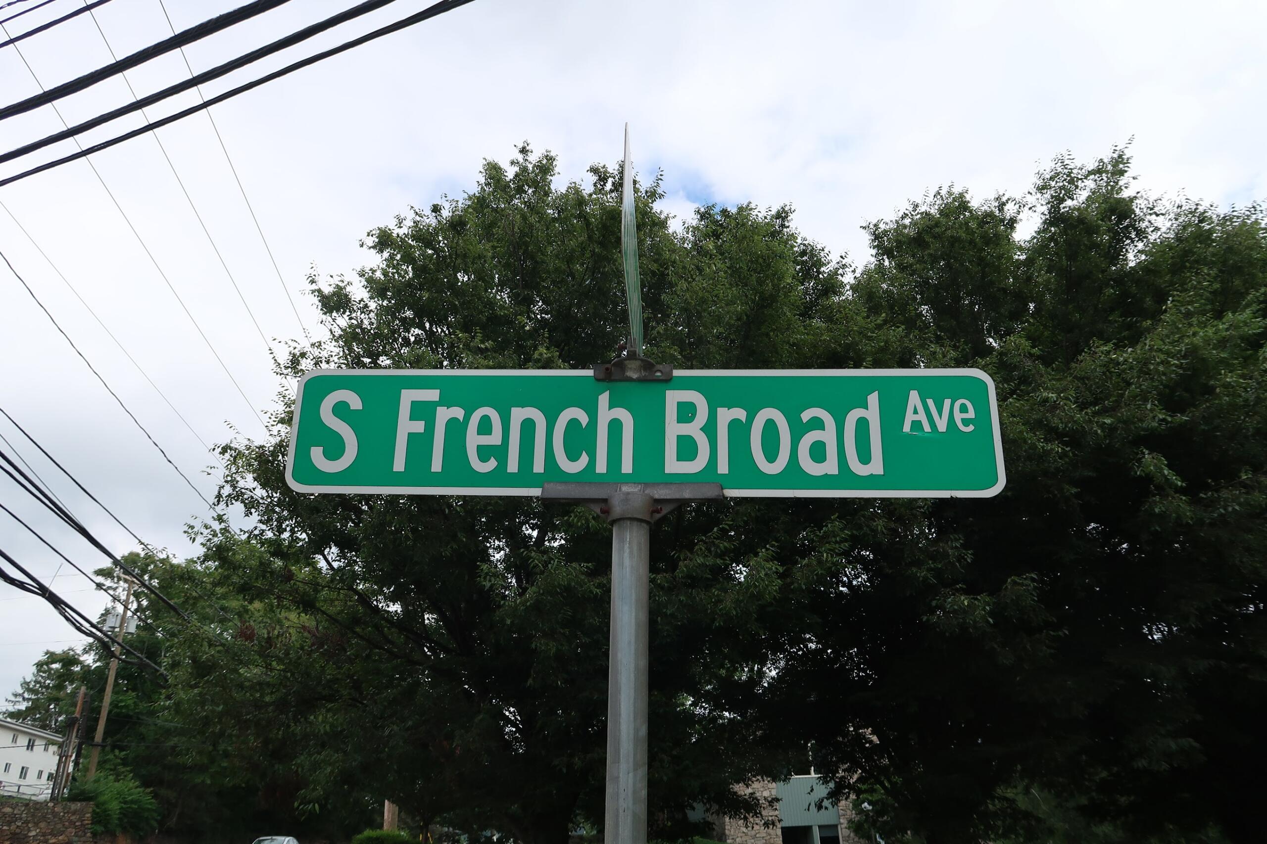 Green street sign reading "S French Broad Ave" with trees and power lines in the background.