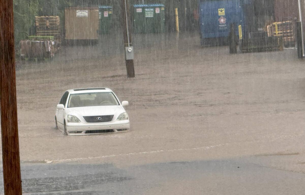 White car partially submerged in floodwaters with heavy rainfall and distant storage containers in the background; scene of severe flooding and rising water levels.