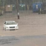 White car partially submerged in floodwaters with heavy rainfall and distant storage containers in the background; scene of severe flooding and rising water levels.
