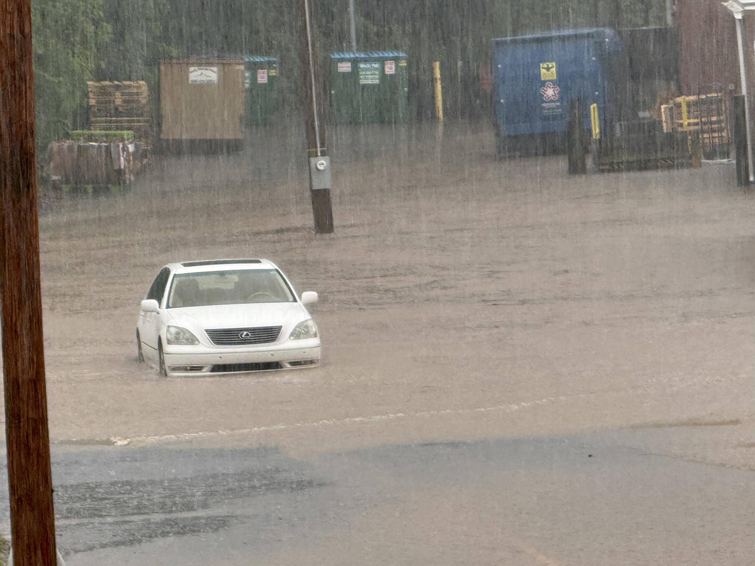 White car partially submerged in floodwaters with heavy rainfall and distant storage containers in the background; scene of severe flooding and rising water levels.