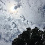 Bright sky with scattered white clouds and sunlight peeking through, above a dark treeline at the bottom of the frame.
