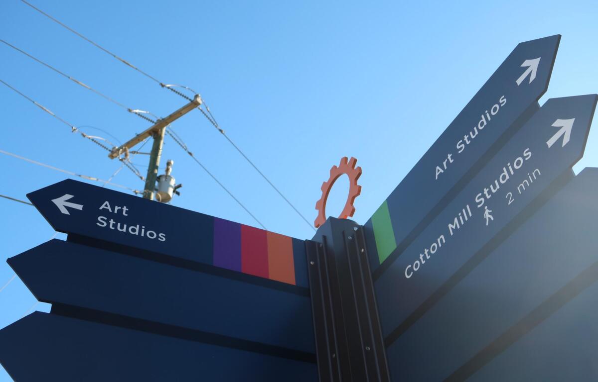 Signpost with blue panels pointing to Art Studios, arrows indicating directions, set against a bright blue sky and colorful striped base.