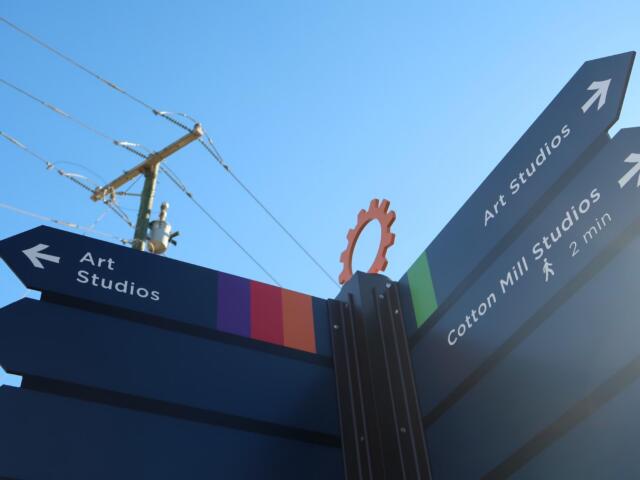 Signpost with blue panels pointing to Art Studios, arrows indicating directions, set against a bright blue sky and colorful striped base.