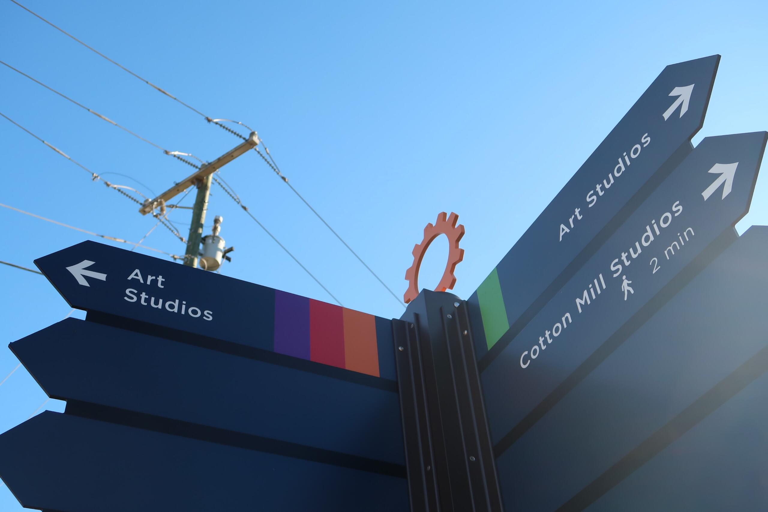 Signpost with blue panels pointing to Art Studios, arrows indicating directions, set against a bright blue sky and colorful striped base.