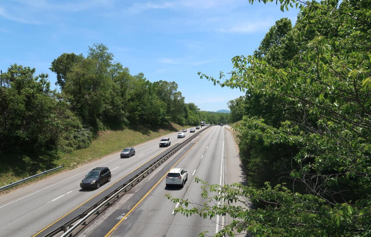 Highway with multiple lanes bordered by green trees under a clear blue sky; several cars travel in both directions.