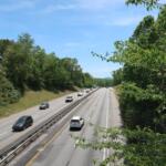 Highway with multiple lanes bordered by green trees under a clear blue sky; several cars travel in both directions.