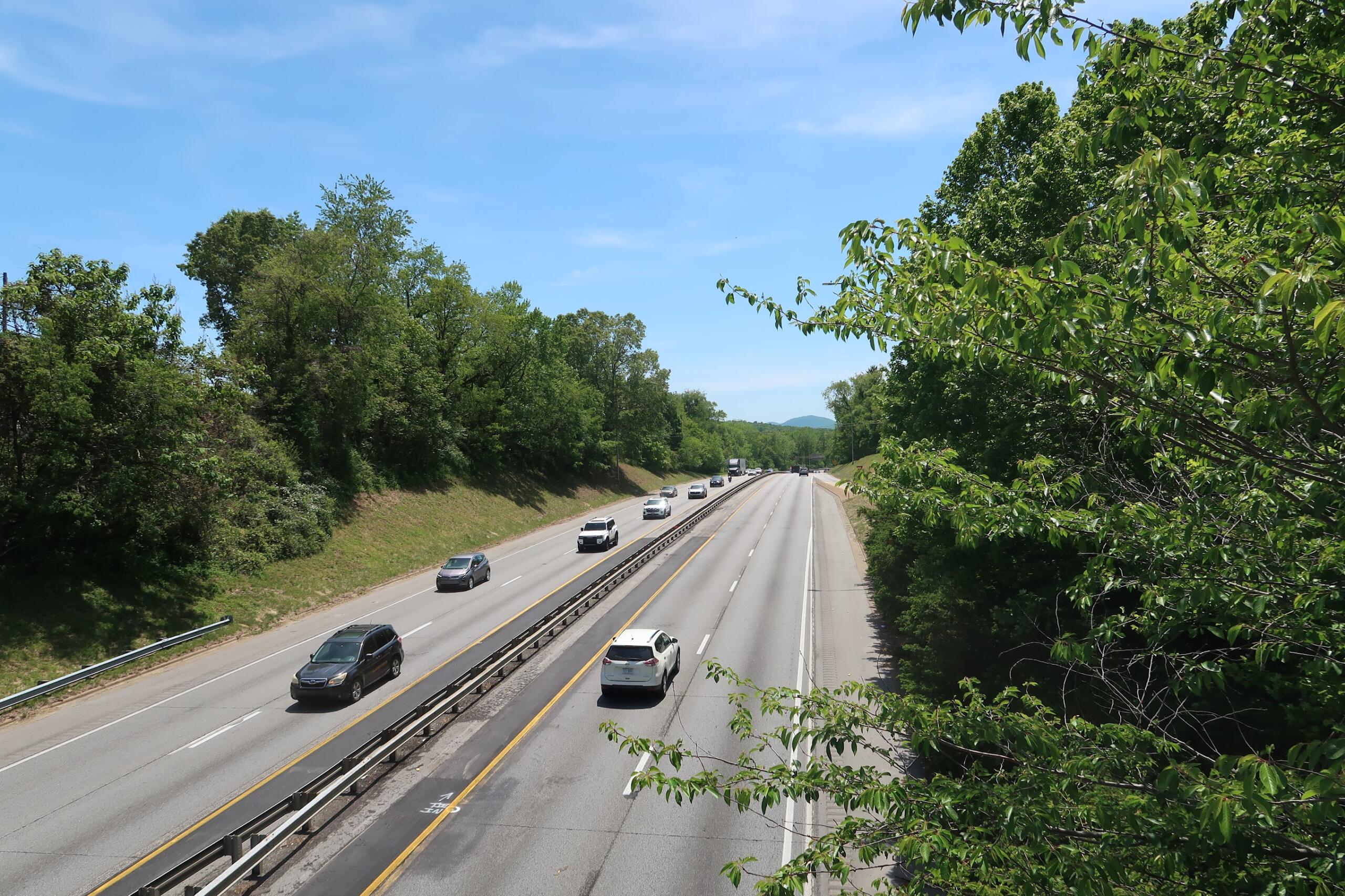 Highway with multiple lanes bordered by green trees under a clear blue sky; several cars travel in both directions.