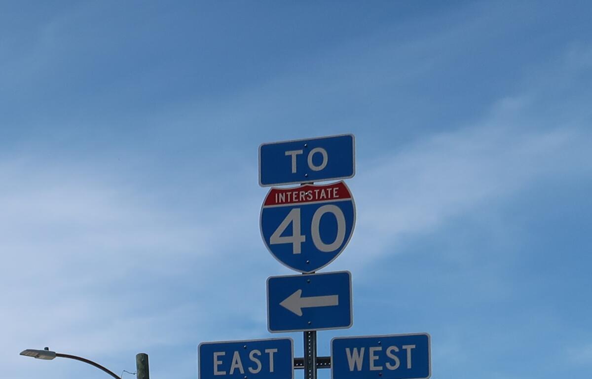 Blue highway sign cluster: 'TO' above a red-arched Interstate 40 shield with large 40, arrows pointing left for directions (east/west signs visible) on a clear sky backdrop.