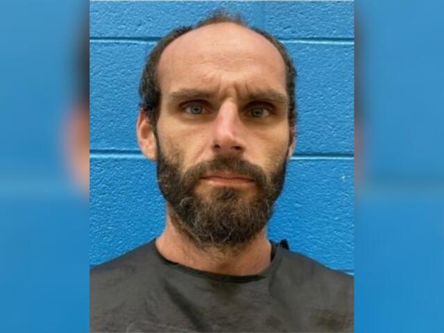 Close-up portrait of a bearded man with a neutral expression in front of a blue brick wall.