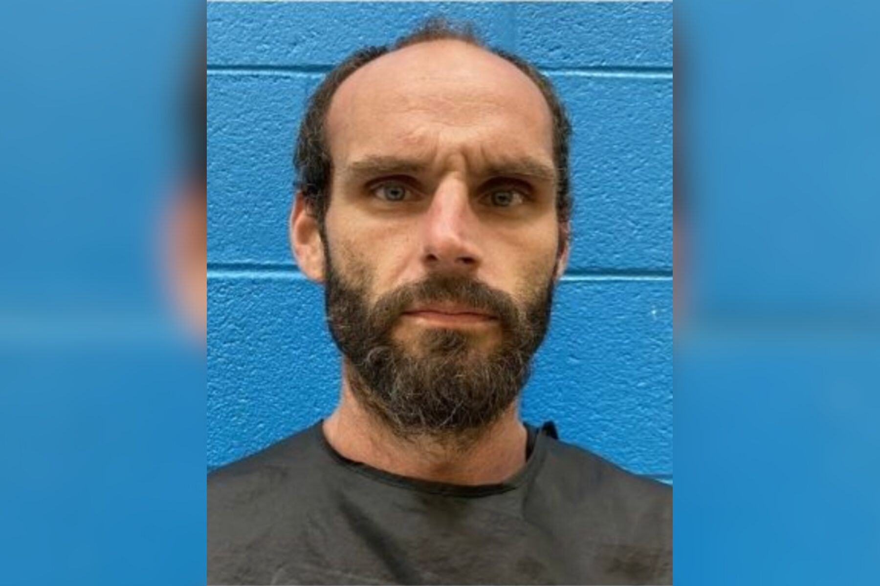 Close-up portrait of a bearded man with a neutral expression in front of a blue brick wall.