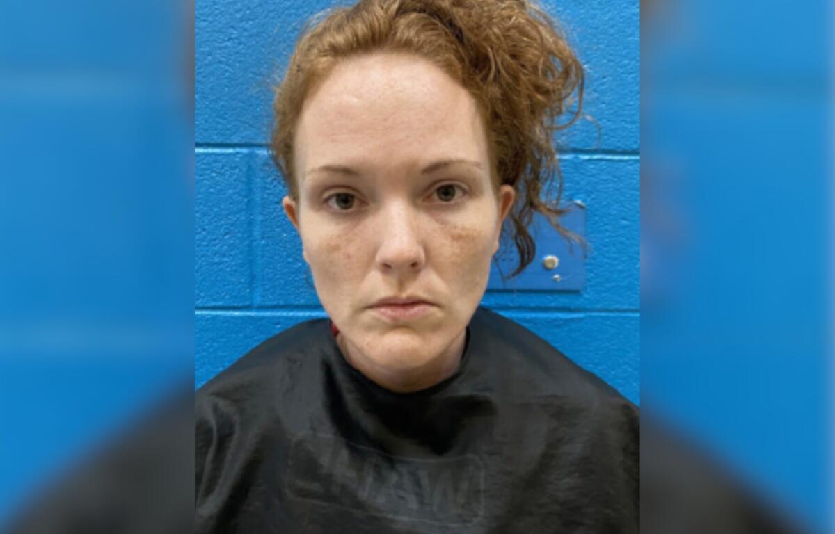 Mugshot-style portrait of a woman with red hair against a blue cinder-block wall, wearing a dark shirt, with facial redness and minor bruising on the cheeks and chin.
