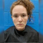 Mugshot-style portrait of a woman with red hair against a blue cinder-block wall, wearing a dark shirt, with facial redness and minor bruising on the cheeks and chin.