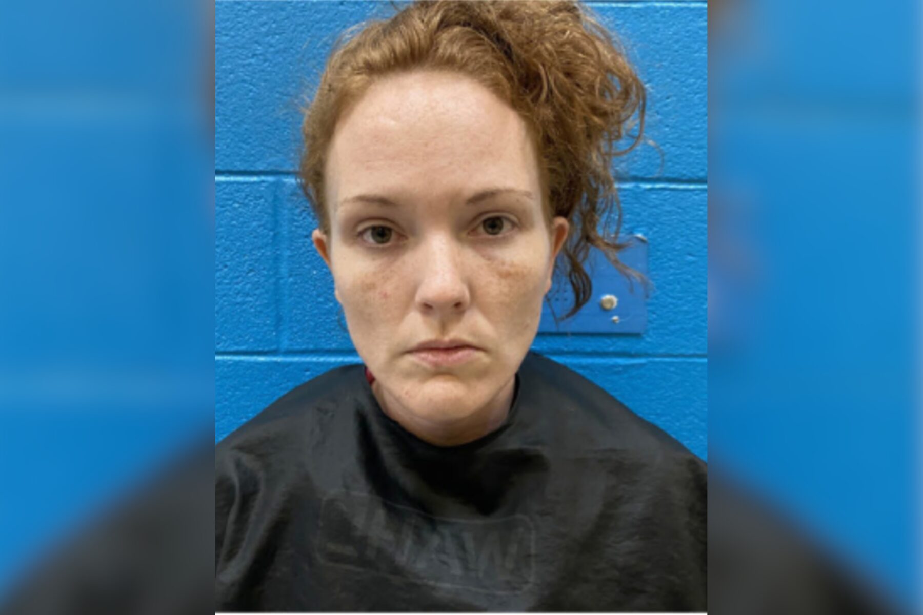 Mugshot-style portrait of a woman with red hair against a blue cinder-block wall, wearing a dark shirt, with facial redness and minor bruising on the cheeks and chin.