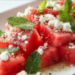 Sliced watermelon topped with crumbled feta, fresh mint, and cracked pepper on a white plate.
