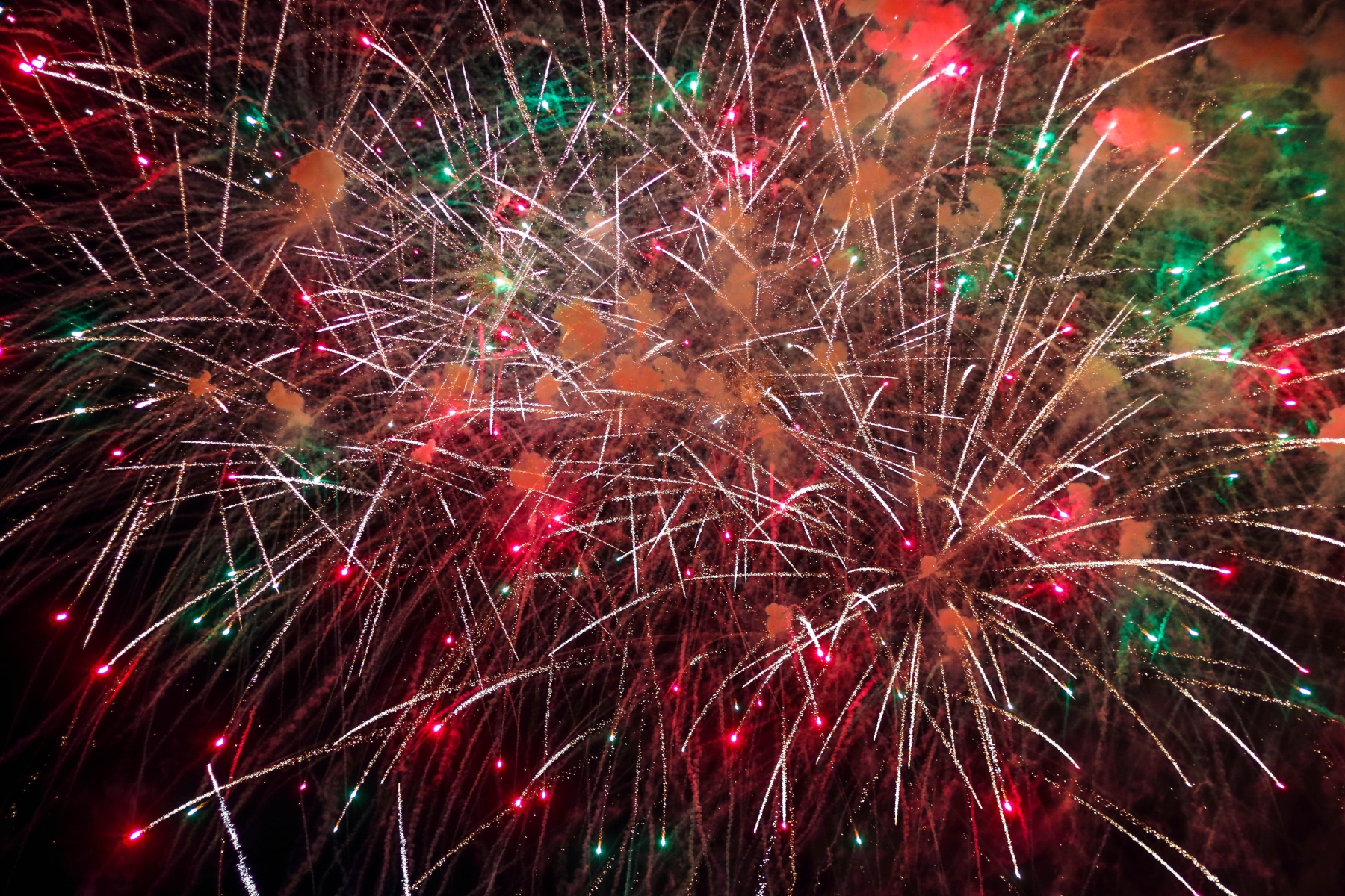 Fireworks explode in a dark sky, white streaks crisscrossing with red and green sparks and smoke.