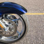 Close-up of a blue motorcycle front fender with decorative pinstripes, chrome forks, and a perforated brake disc.
