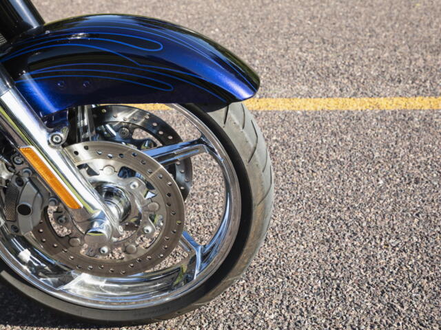 Close-up of a blue motorcycle front fender with decorative pinstripes, chrome forks, and a perforated brake disc.