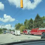 Red fire trucks and emergency vehicles line a city street on a sunny day with green trees and traffic lights overhead
