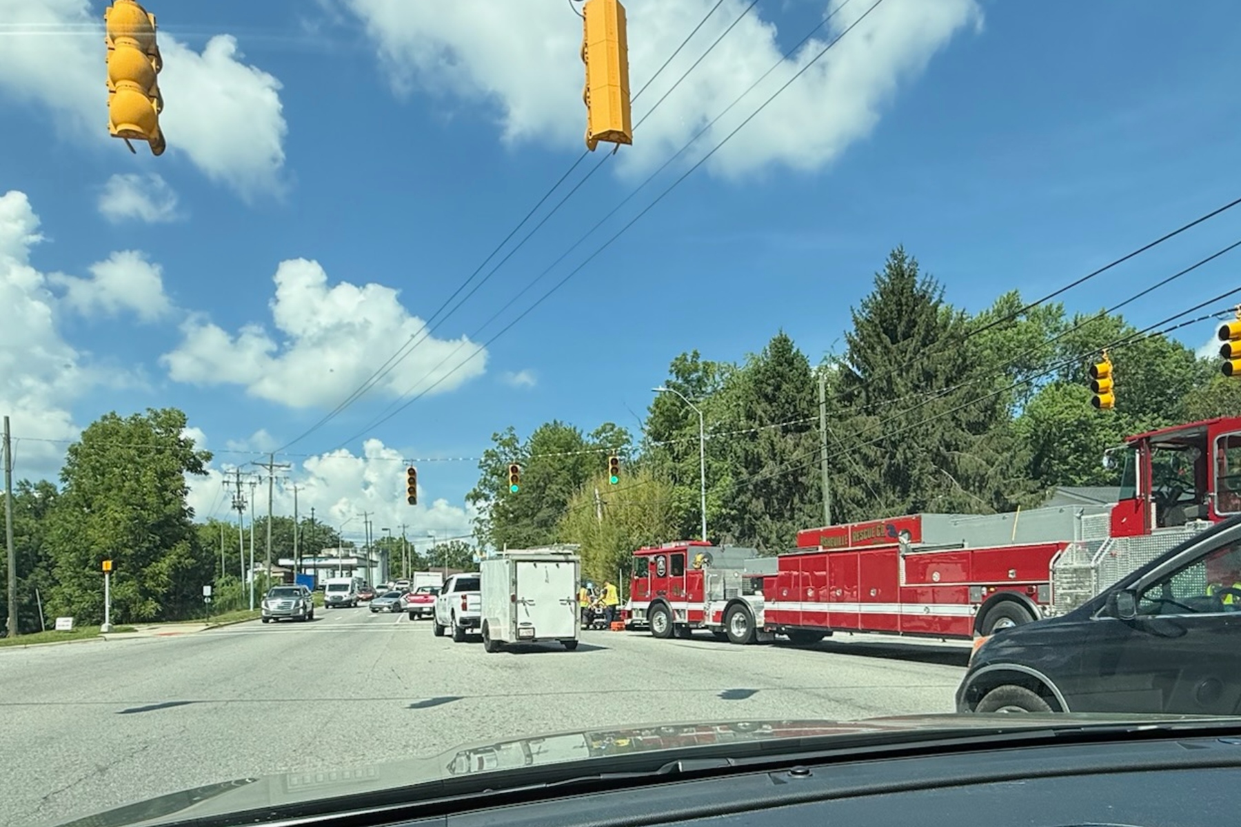 Red fire trucks and emergency vehicles line a city street on a sunny day with green trees and traffic lights overhead