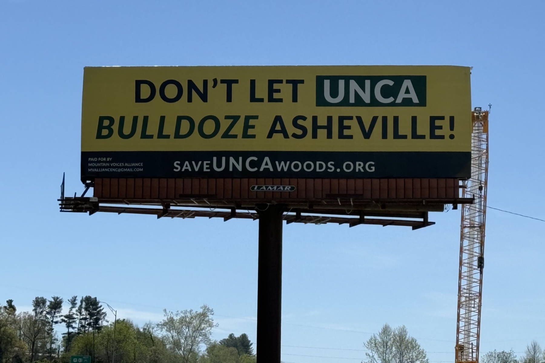 A "Save the Woods" billboard stands along Future I-26 in Asheville, highlighting community opposition to potential development of UNC Asheville's South Campus woods.