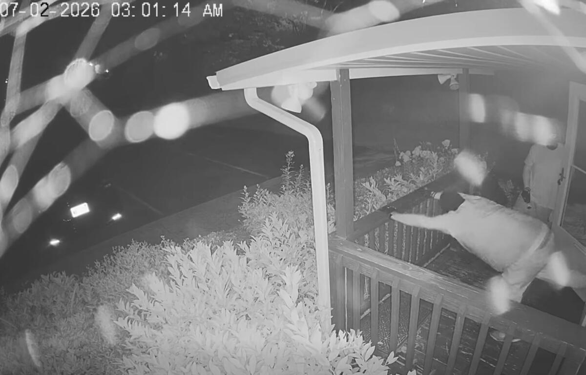 Night-vision porch scene: a person in light clothing reaches toward the door while another person stands on the porch; bushes and railing visible; timestamp 07-02-2026 03:01:14 AM.