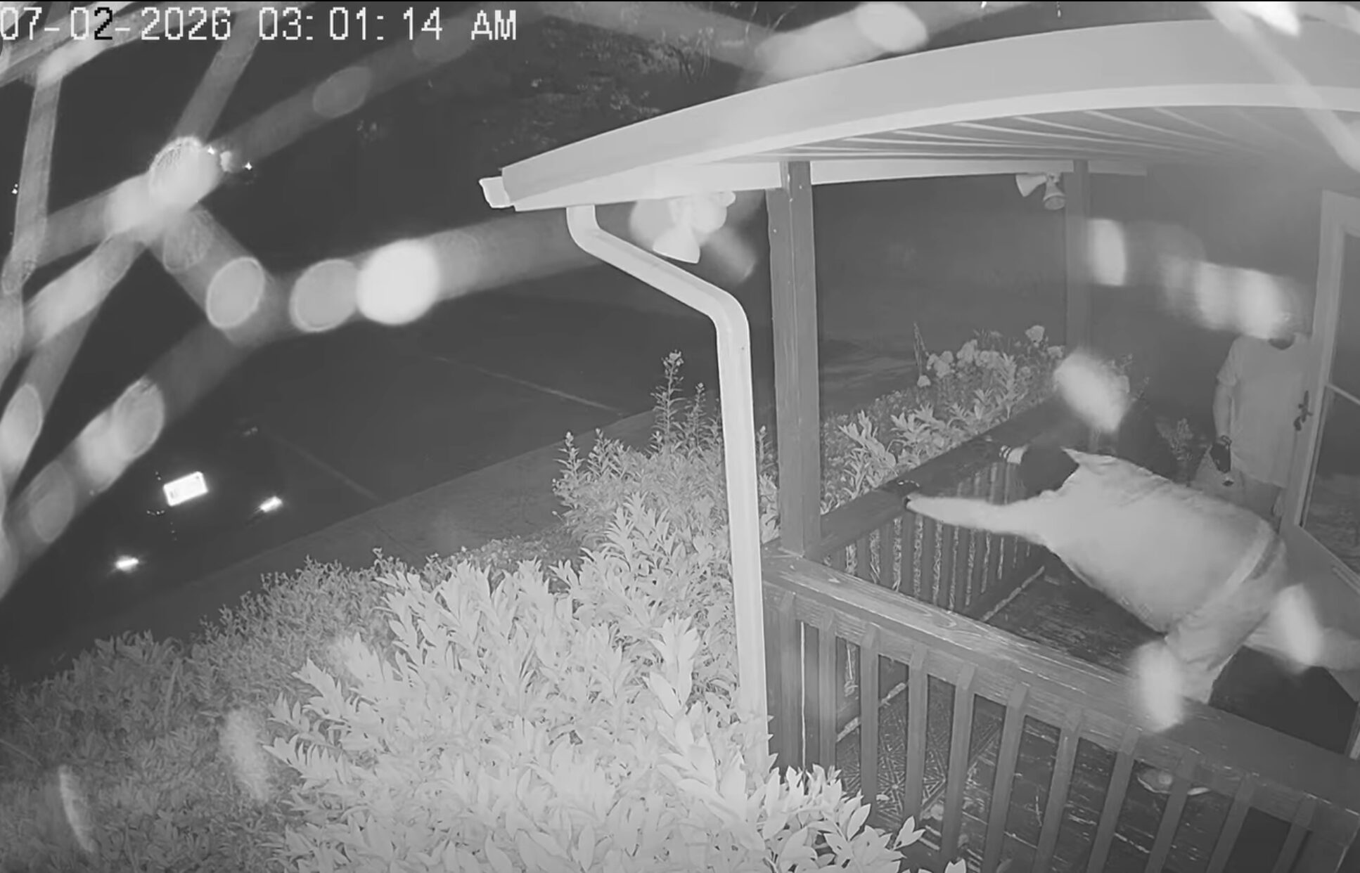 Night-vision porch scene: a person in light clothing reaches toward the door while another person stands on the porch; bushes and railing visible; timestamp 07-02-2026 03:01:14 AM.